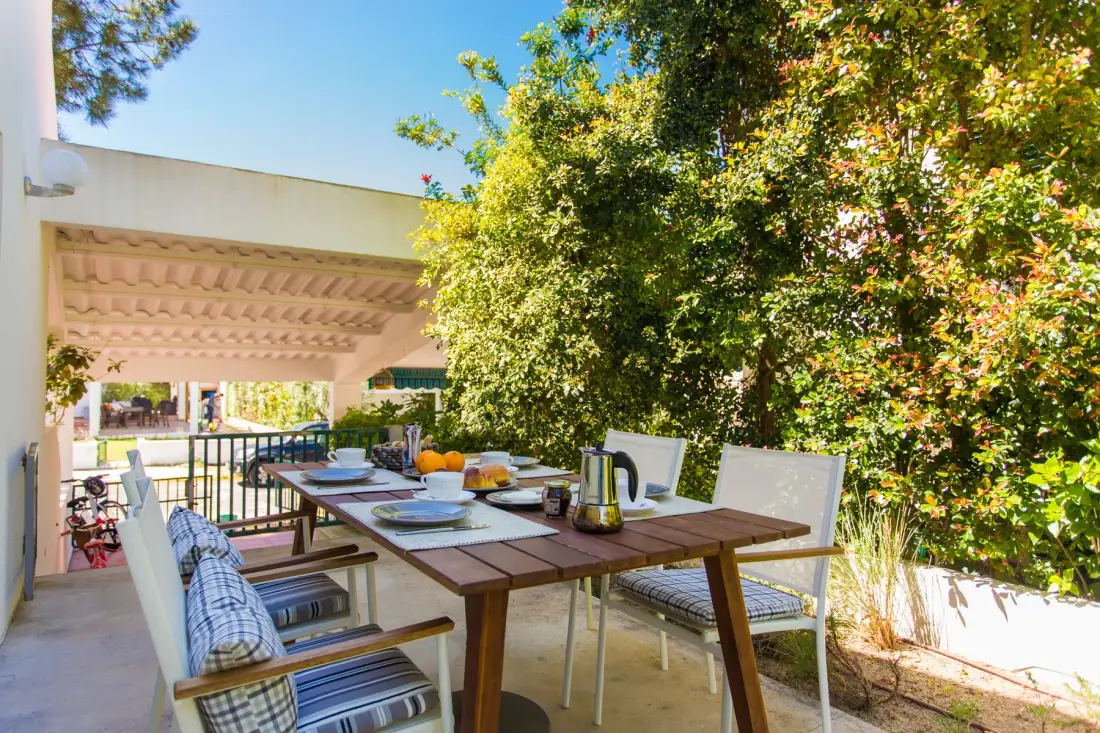 Zona de comedor al aire libre con una mesa de madera y asientos bajo un árbol frondoso.