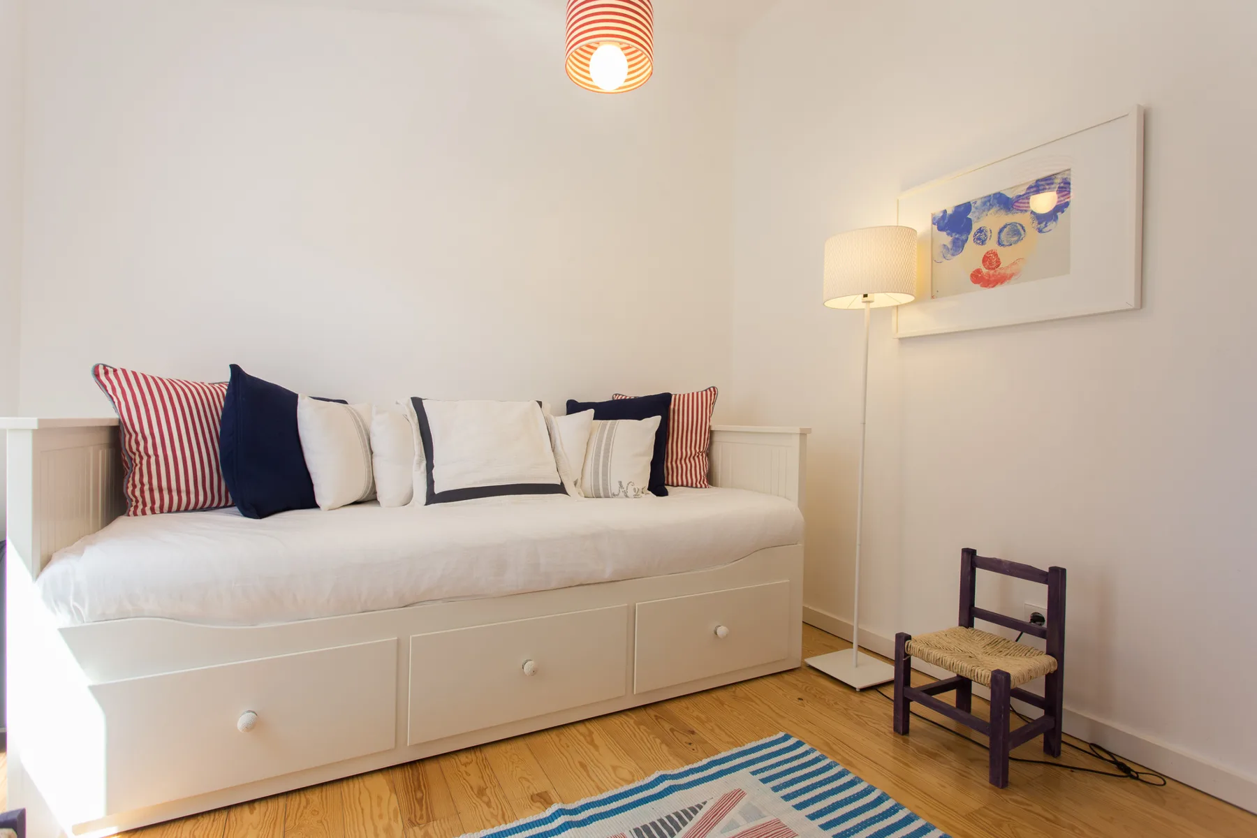 Un dormitorio luminoso con una cama blanca, cojines estampados y una silla pequeña.