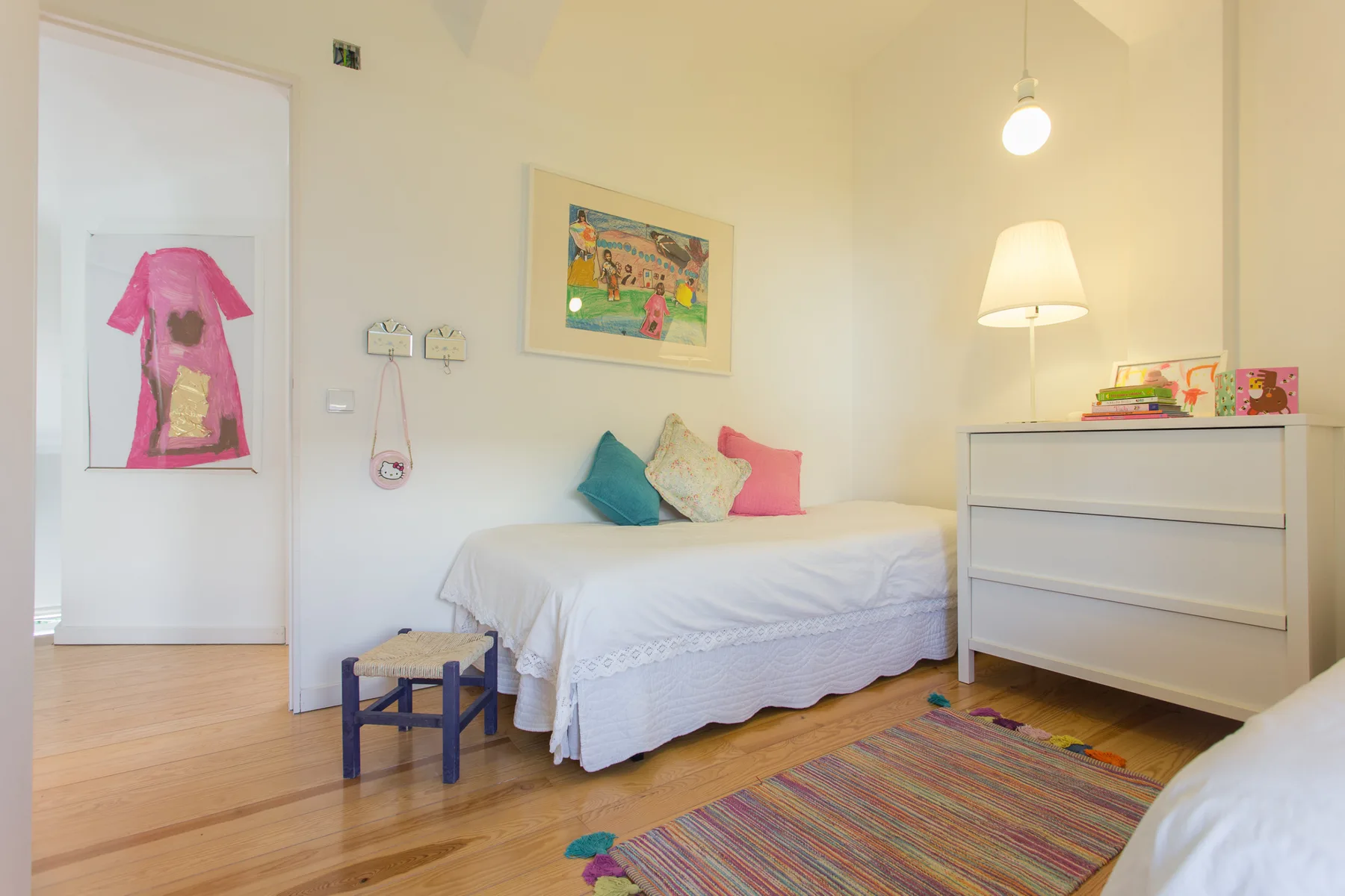 Un dormitorio luminoso con una cama, una cómoda y obras de arte coloridas.