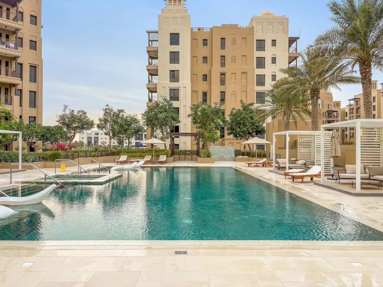 An outdoor swimming pool is surrounded by lounge seating, palm trees, and apartment buildings.