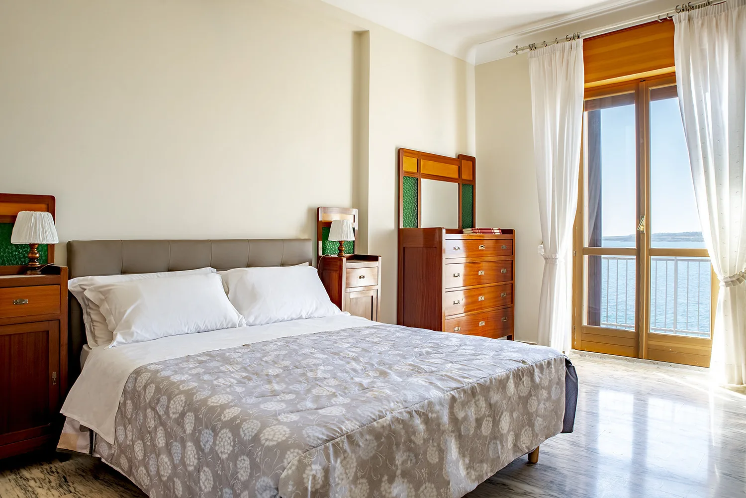 Un dormitorio con una cama doble, una cómoda de madera y una ventana alta con vistas al mar.