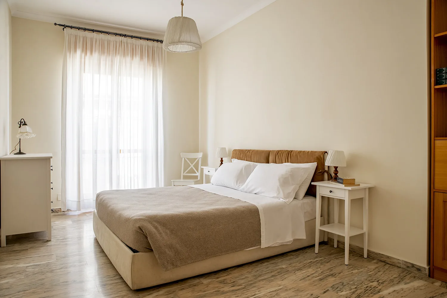 Un dormitorio luminoso con una cama junto a una ventana alta con cortinas transparentes.