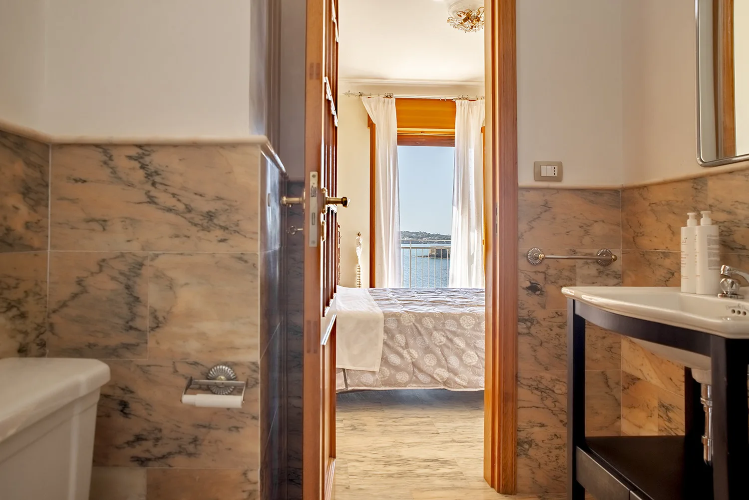 La puerta del baño da a un dormitorio con una cama y una ventana luminosa.