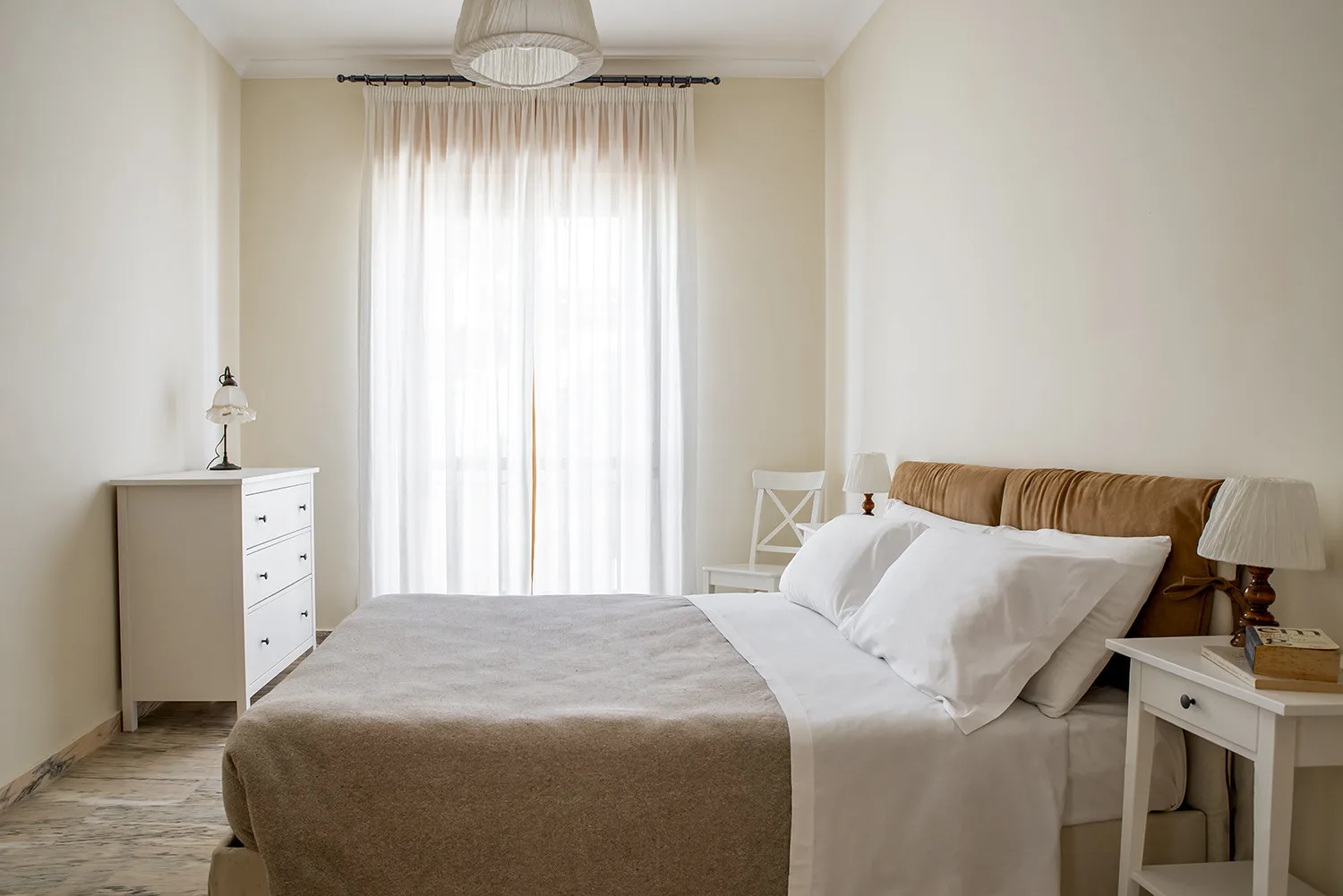 Un dormitorio luminoso con una cama bien hecha y cortinas translúcidas en la ventana.