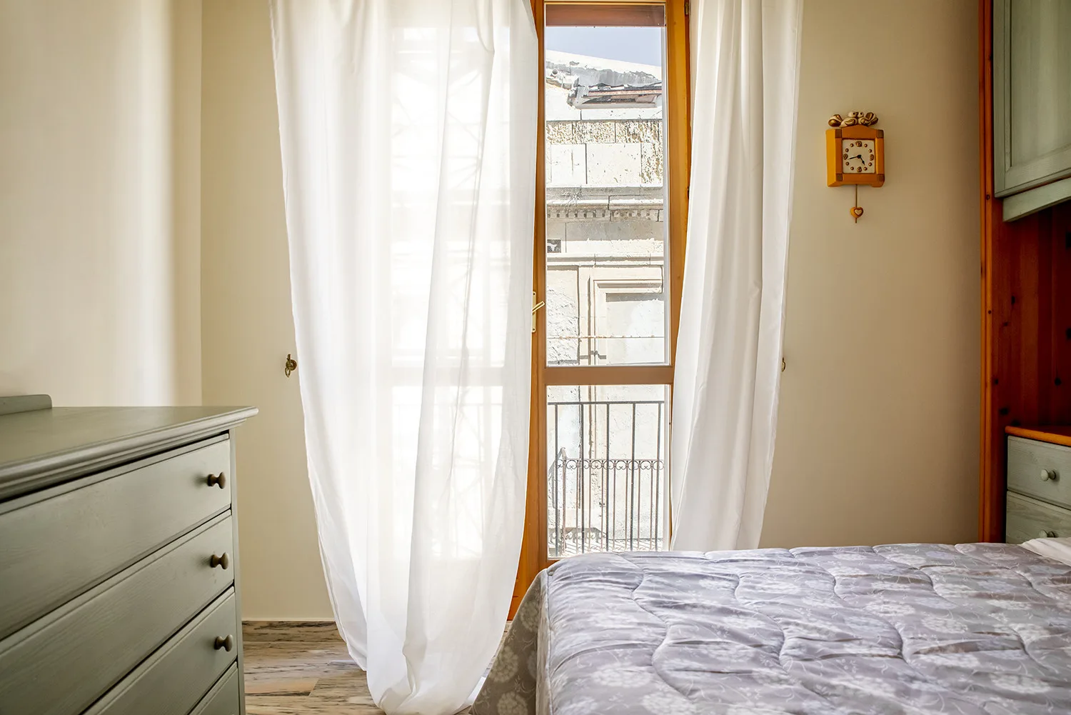 Un dormitorio con una cama junto a una ventana alta enmarcada por cortinas blancas.