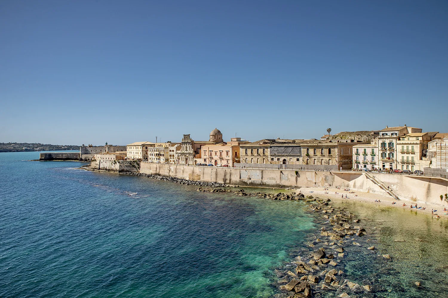 Un pueblo costero se extiende junto a un paseo de piedra y el mar azul claro.