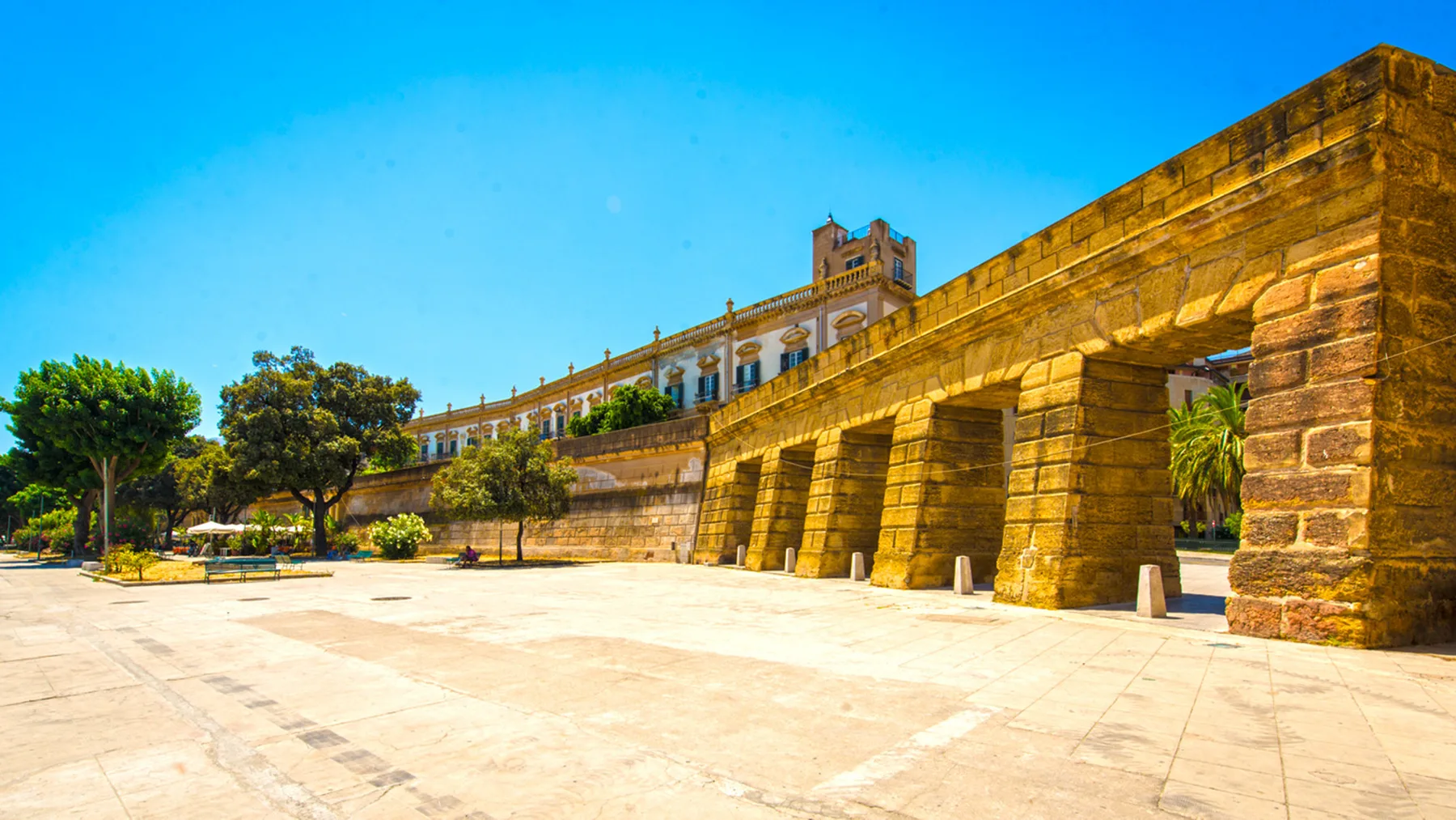 A broad stone plaza beside a long historic-looking masonry wall under a clear blue sky.