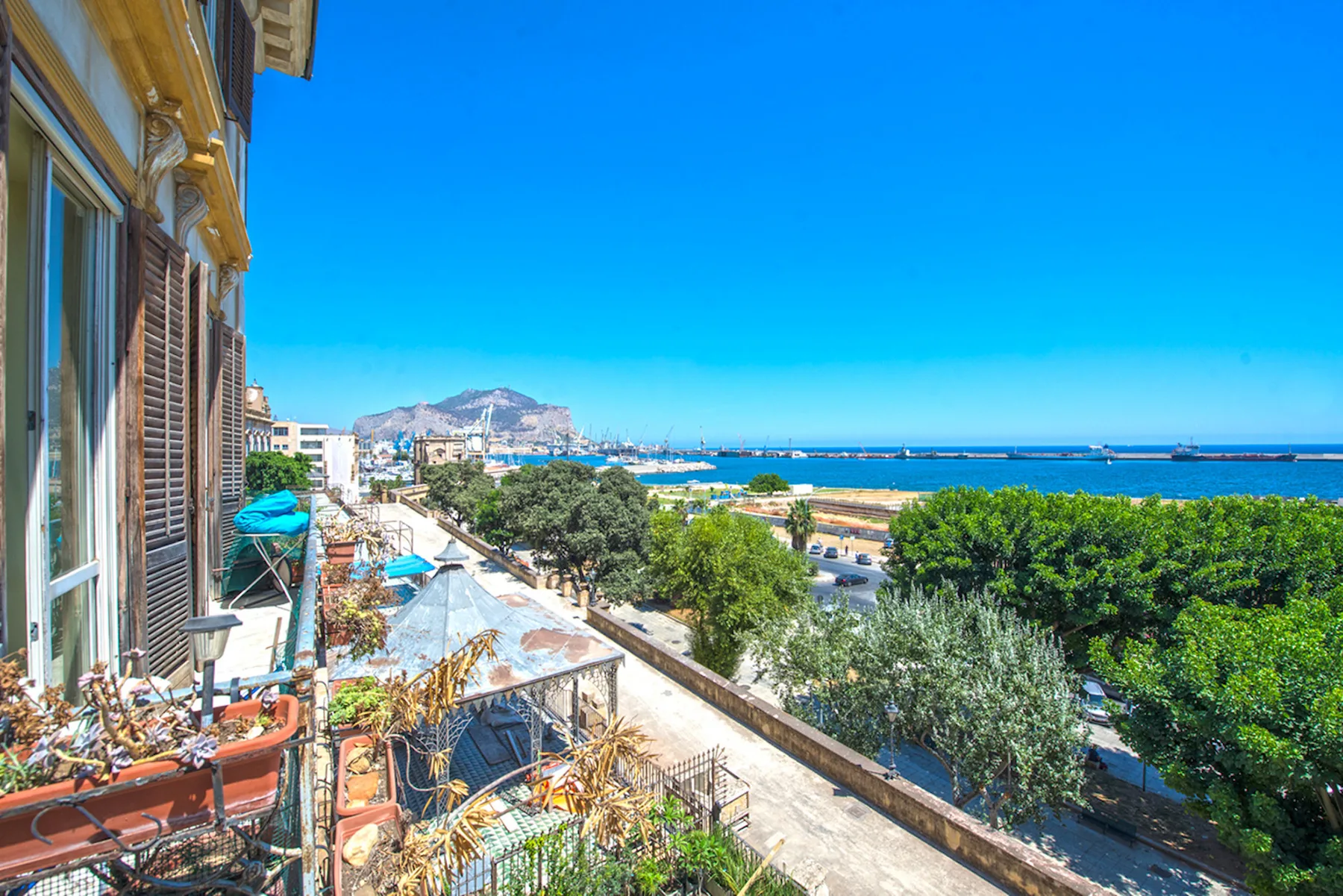 A seaside balcony overlooks a tree-lined promenade, the bay, and a distant mountain.