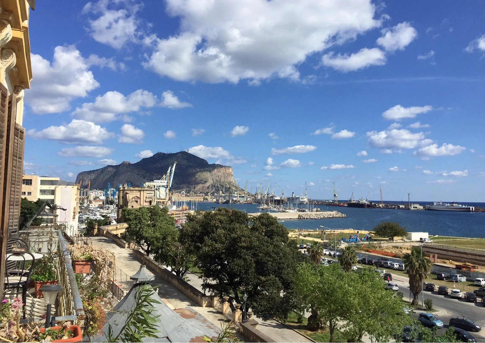 A coastal harbor and marina sit below a mountain under a bright blue sky.