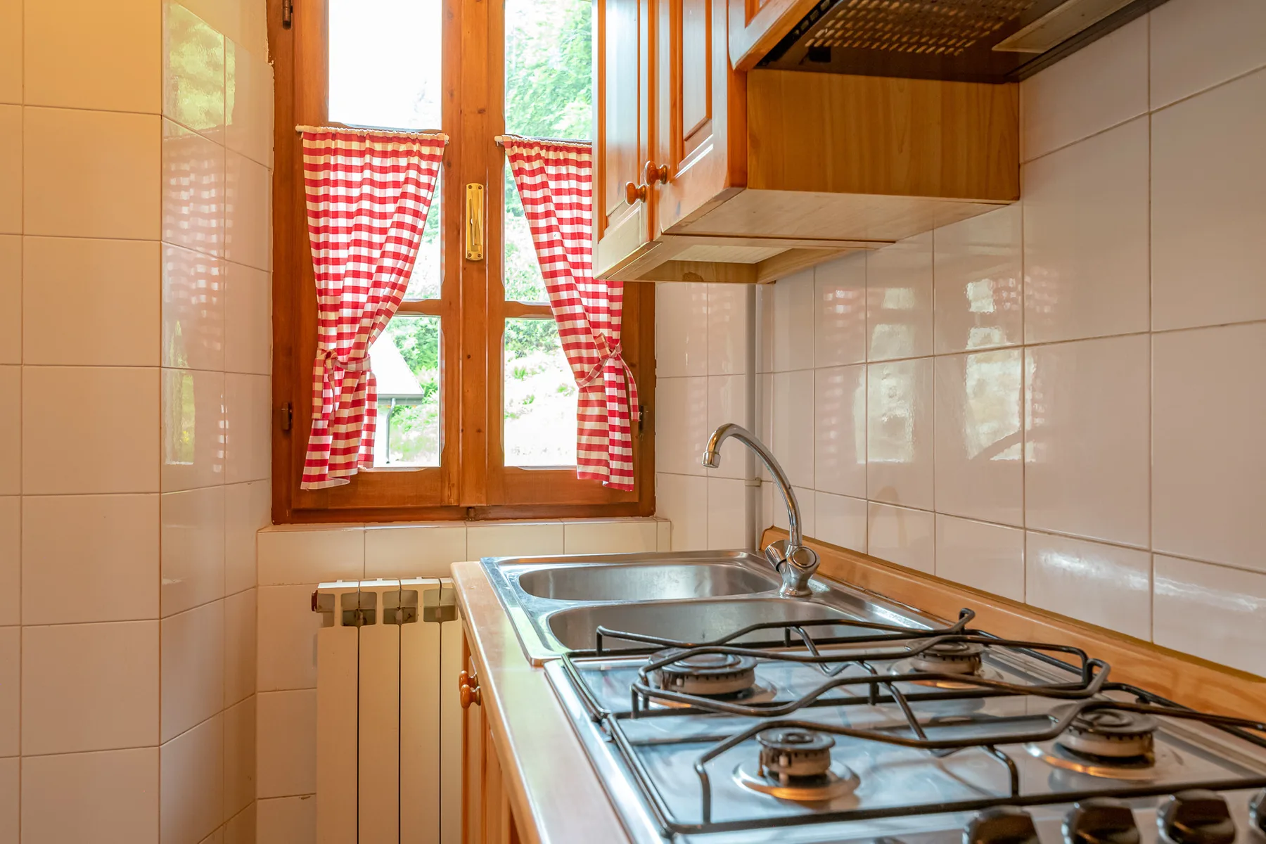 Una cocina alicatada con una placa de gas bajo una ventana con cortinas rojas y blancas.