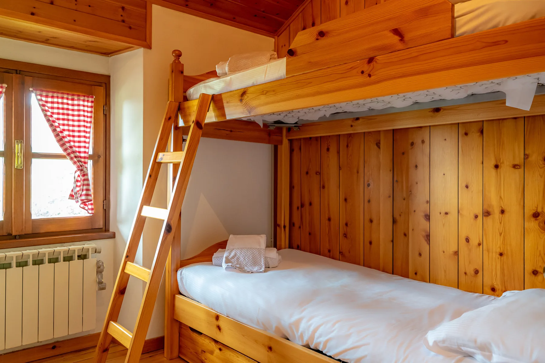 Una litera de madera ocupa un dormitorio pequeño junto a una ventana con cortina a cuadros.