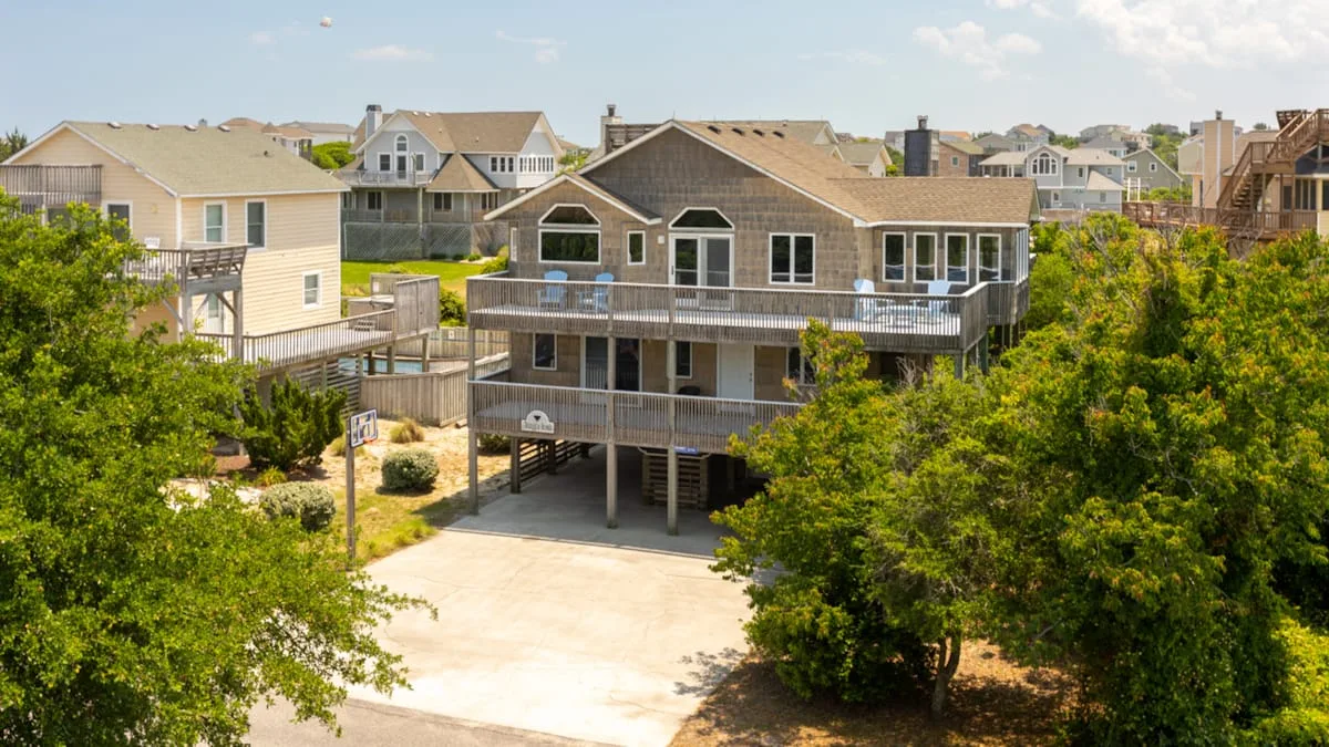 A large elevated house stands among trees beside a broad paved area.