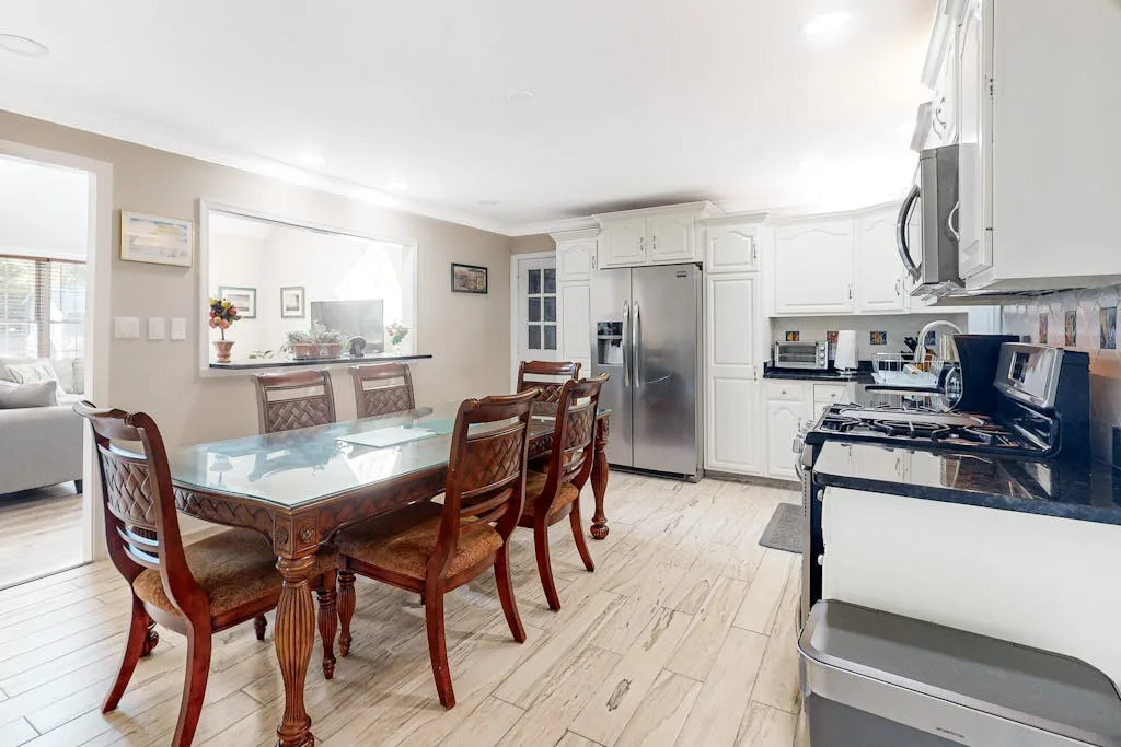 A dining area with a glass-topped table beside a kitchen with white cabinets.