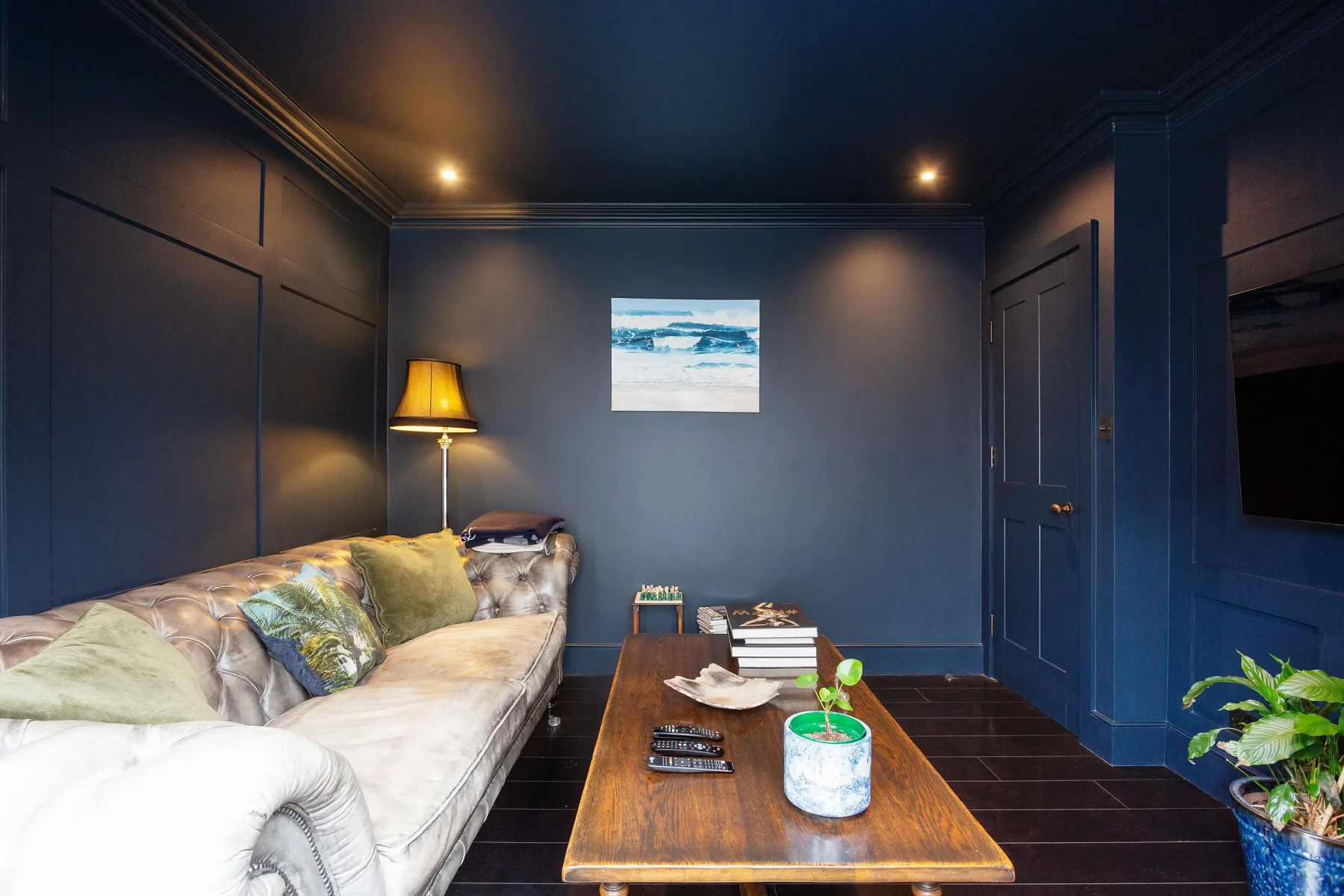 Sala de estar azul oscuro con asientos, una mesa de centro de madera y un paisaje marino enmarcado.