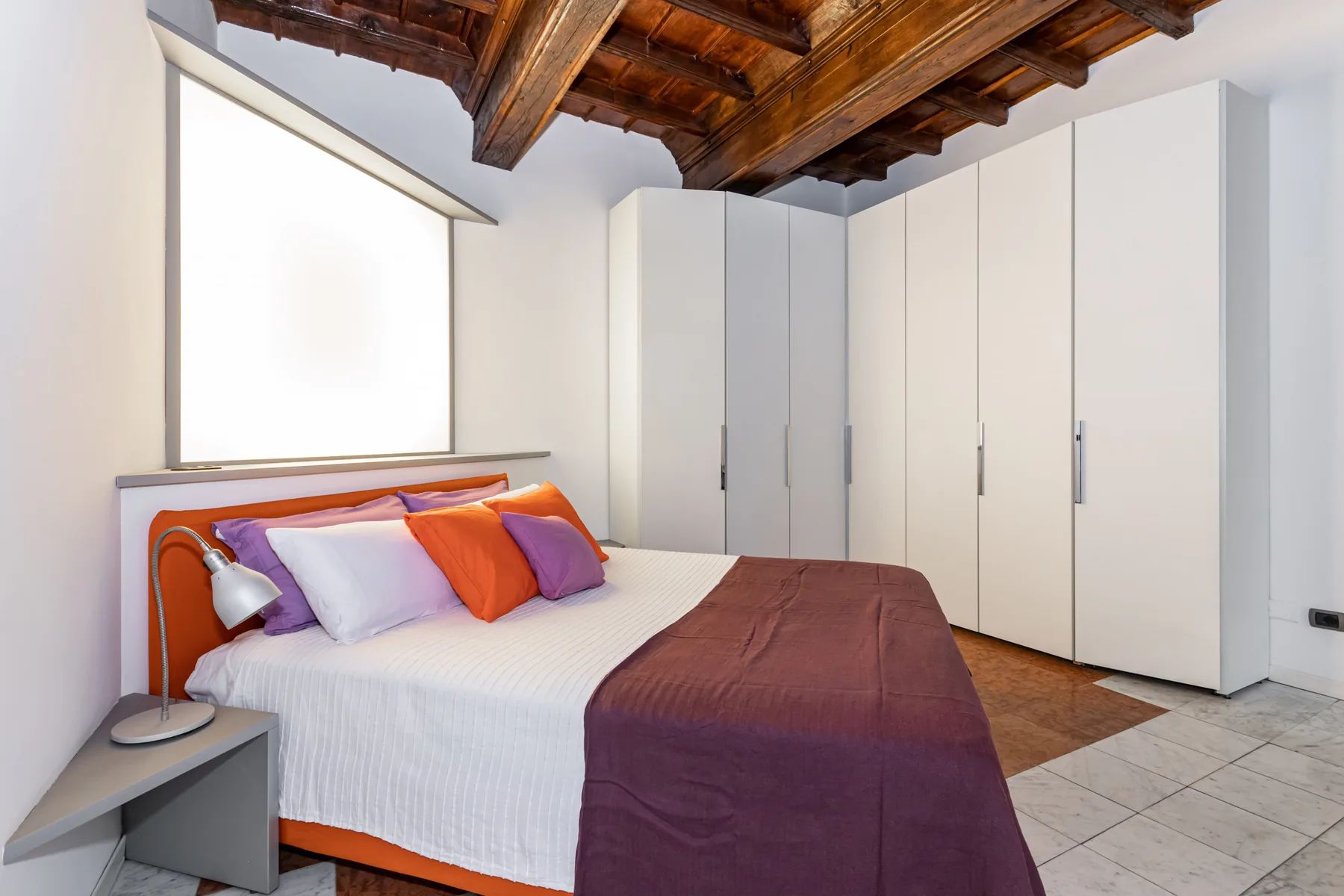 A bedroom with a bed dressed in white and purple bedding beneath exposed wooden ceiling beams.