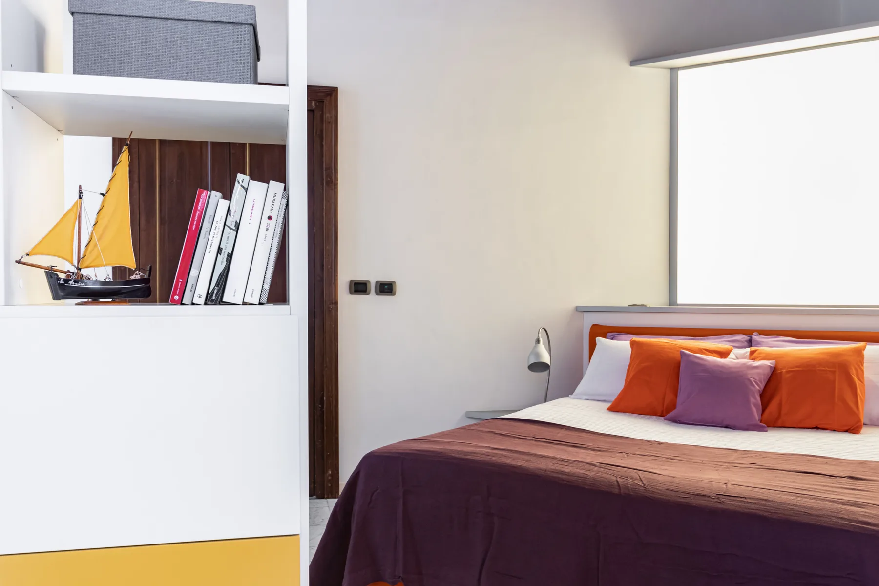 A bedroom with a bed dressed in purple and orange bedding beside built-in shelving.