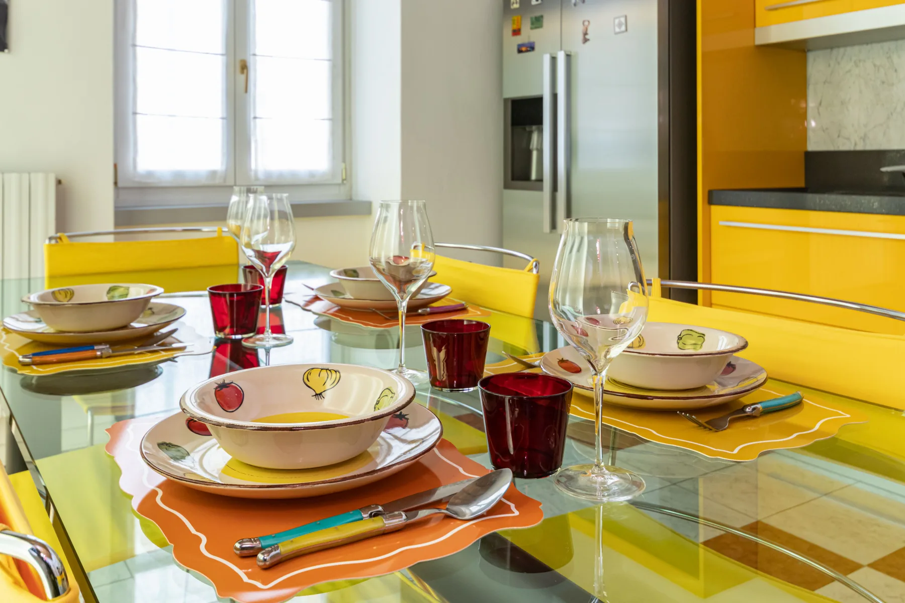 A dining table is set with plates, glasses, and colorful placemats beside a bright kitchen.