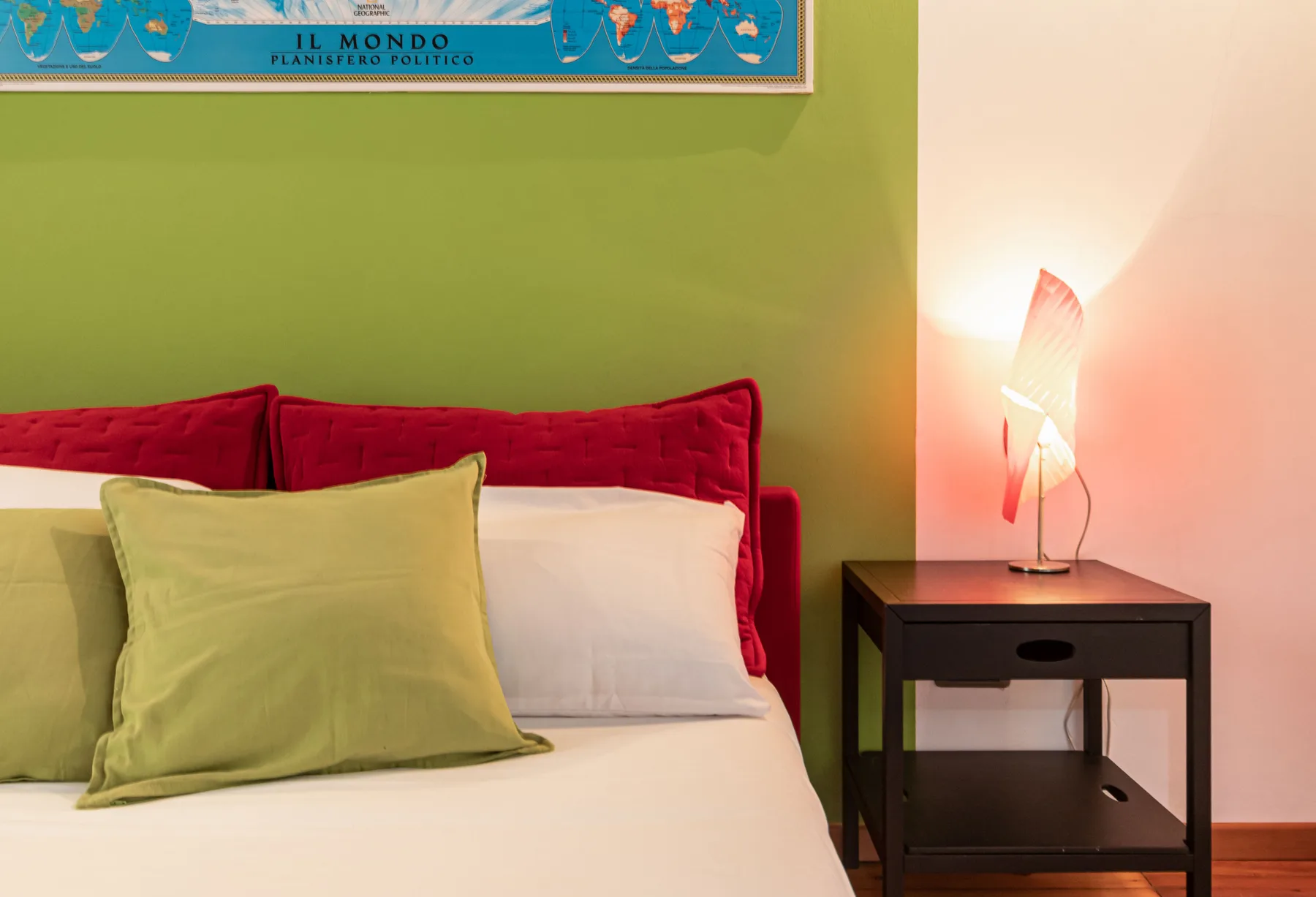 A bed with green and red cushions sits beside a lit bedside table.