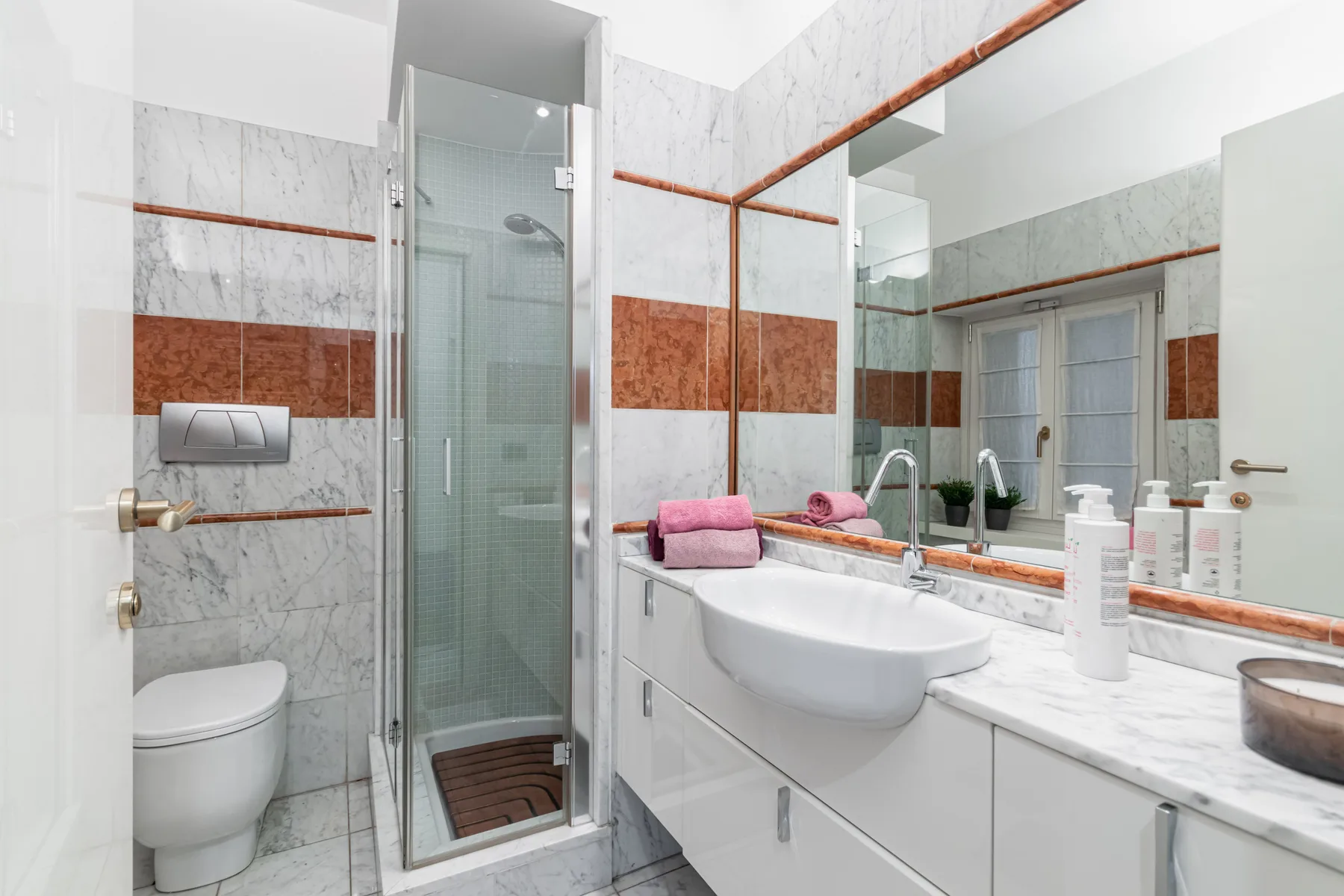 A bright tiled bathroom with a glass shower, toilet, and sink beside a wide mirror.
