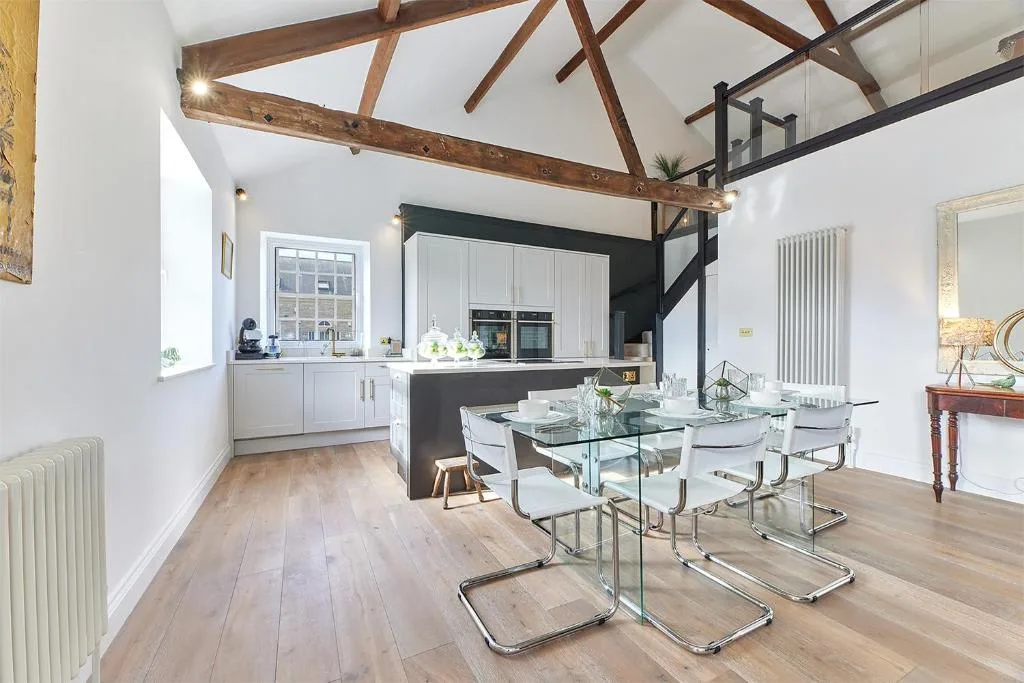 A bright dining area with a glass table, white seating, and an open kitchen.