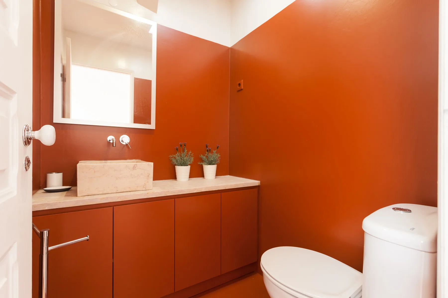 A bathroom with a sink, mirror, and toilet against terracotta-colored walls.
