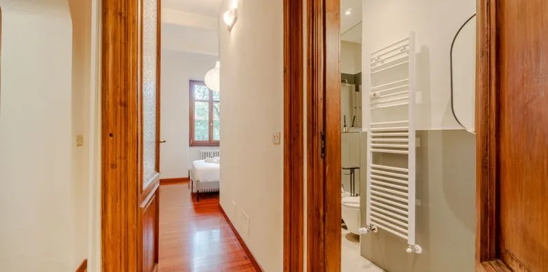 Un pasillo con puertas de madera pasa junto a un baño con un toallero calefactable.