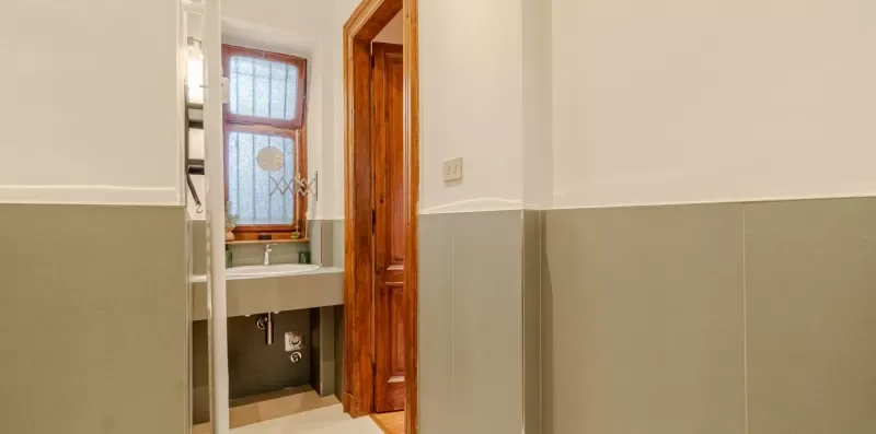 Un baño con azulejos, un lavabo bajo una ventana y una puerta de madera abierta.