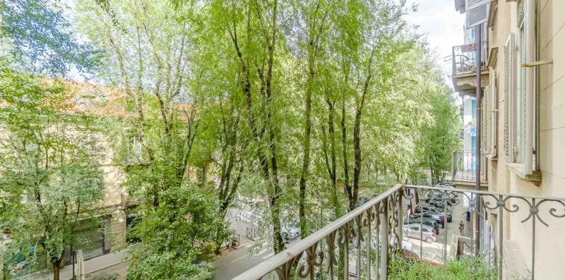 Un balcón da a árboles frondosos y una calle con coches aparcados.