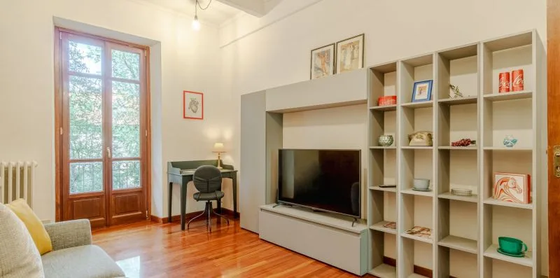 Una sala luminosa con televisor, estanterías abiertas y puertas altas acristaladas.