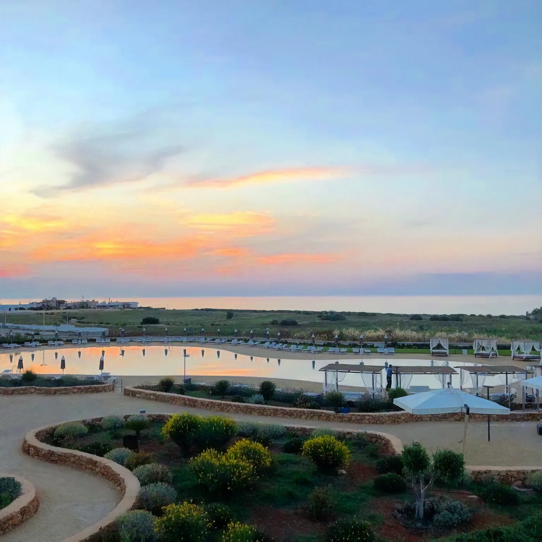 A landscaped outdoor pool area overlooks the sea beneath a colorful sunset sky.