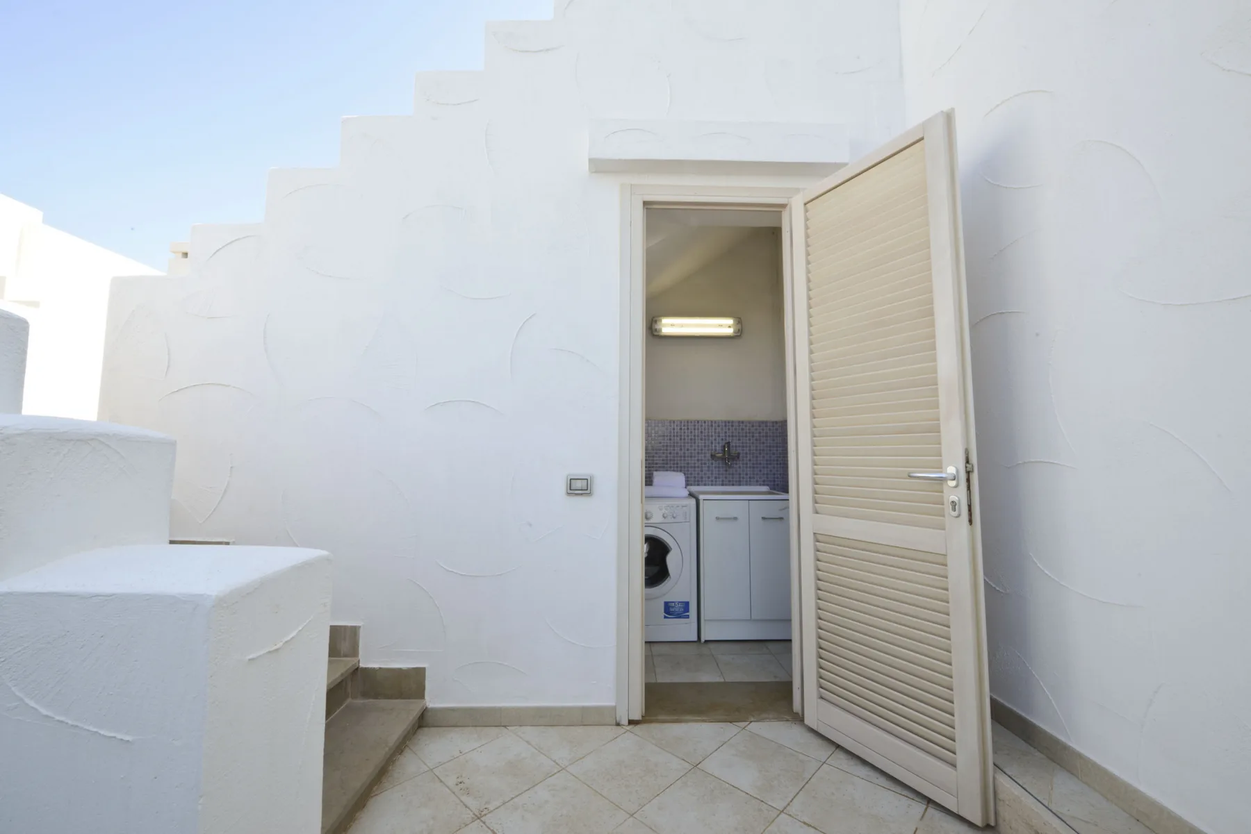 A white patio with steps leads to an open doorway showing a laundry area.