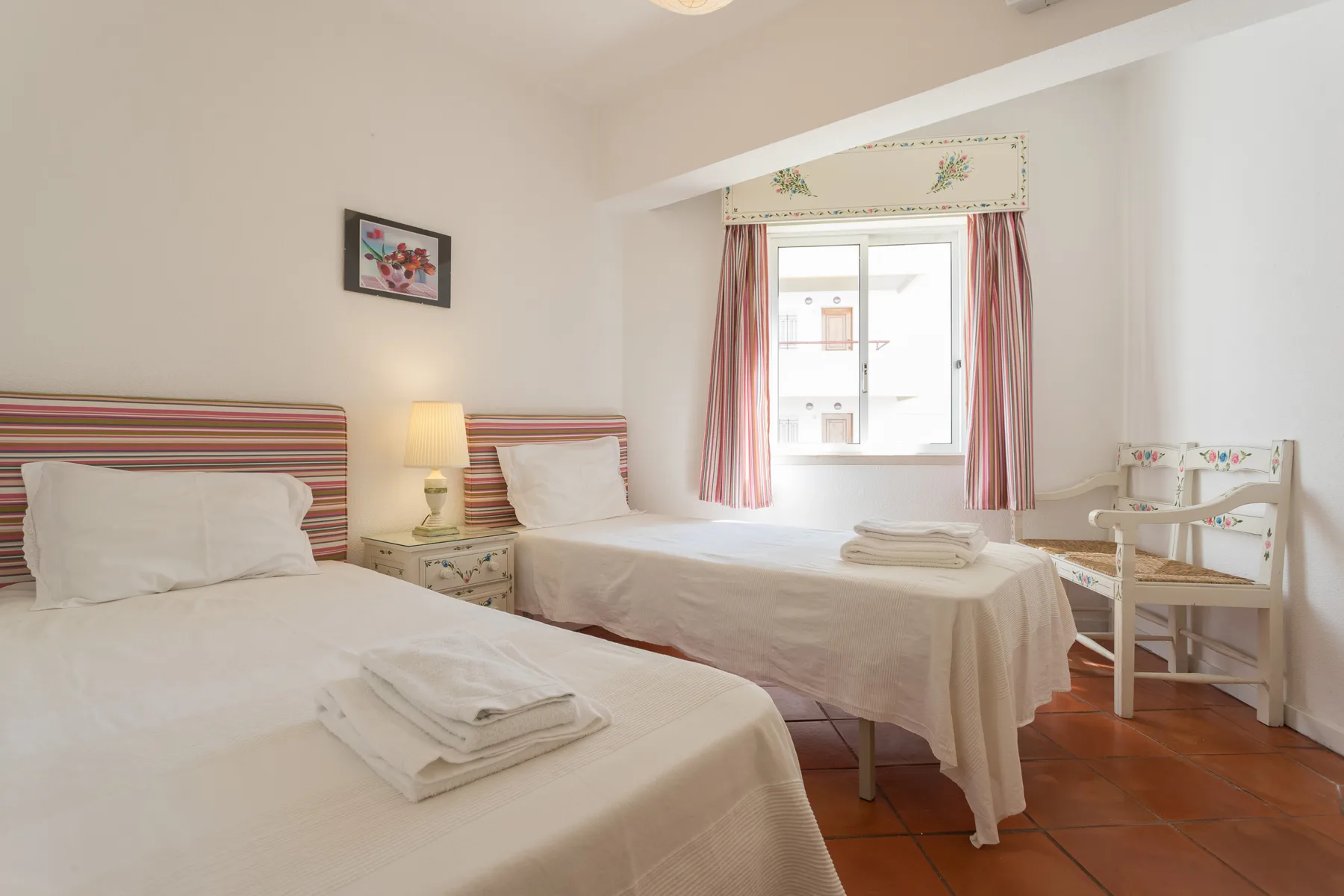A bright bedroom with two beds, bedside tables, and a window.