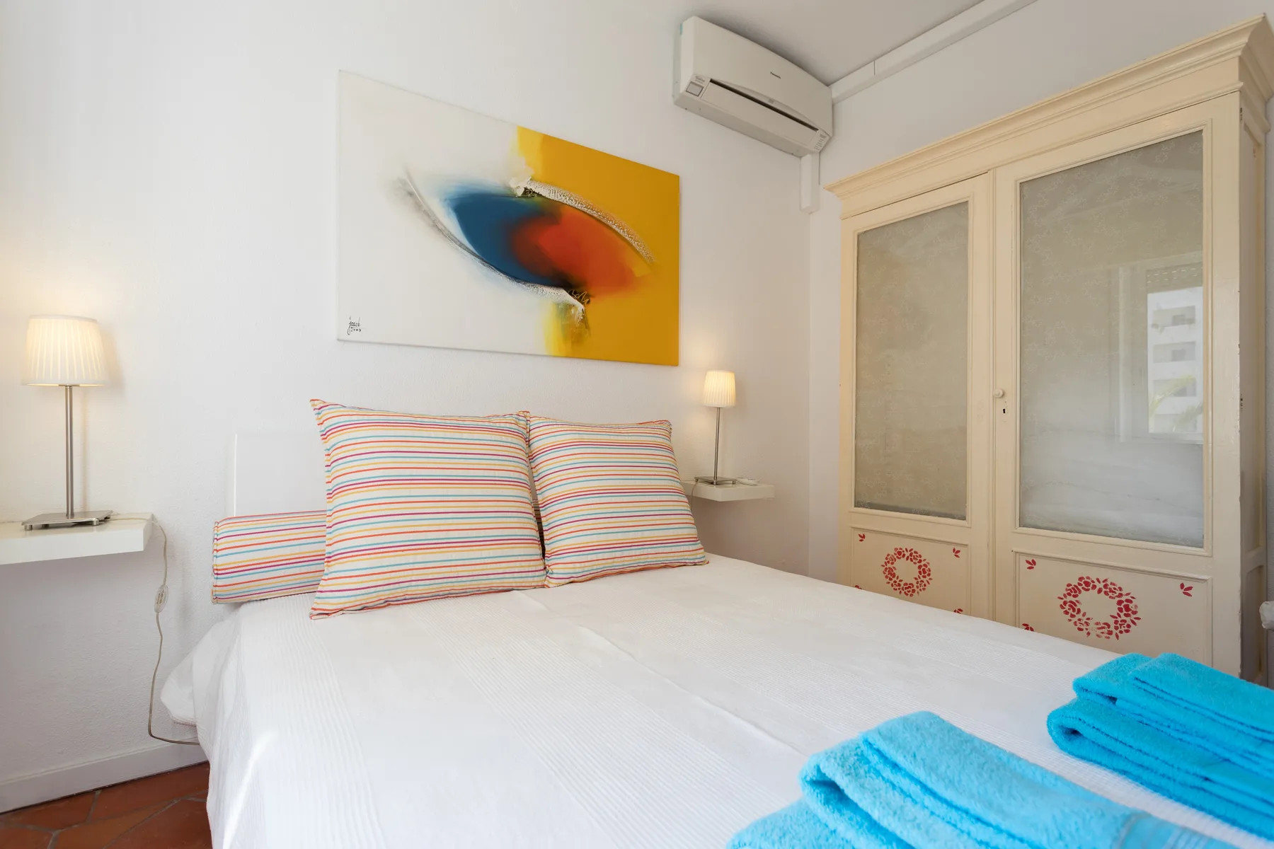 A bedroom with a neatly made bed, striped pillows, and colorful artwork above.