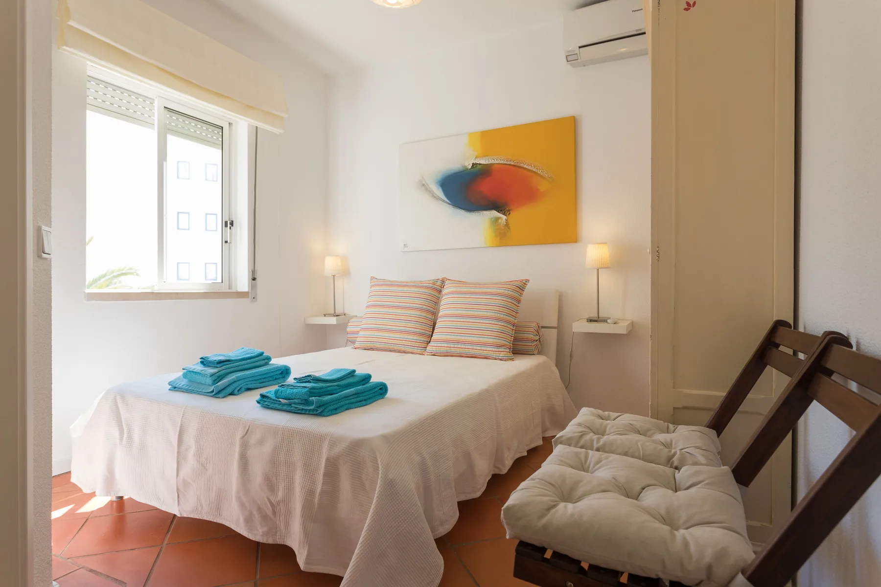 Un dormitorio con una cama, toallas dobladas y una obra colorida sobre el cabecero.