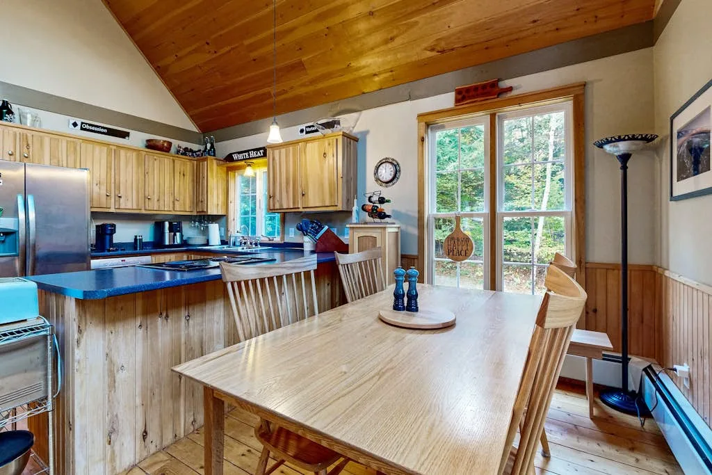 A dining area with a wooden table beside a kitchen and large windows.