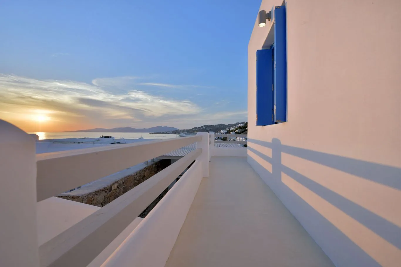 A terrace overlooks the sea at sunset beside a white building with blue shutters.