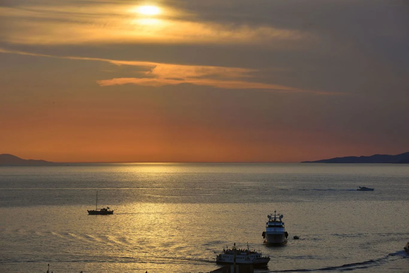 A sunset glows over calm water, with boats near the distant shoreline.
