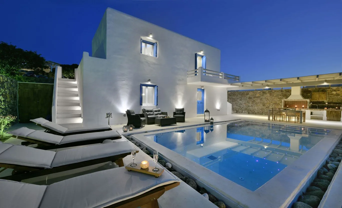 A modern villa exterior with a swimming pool and lounge seating at dusk.