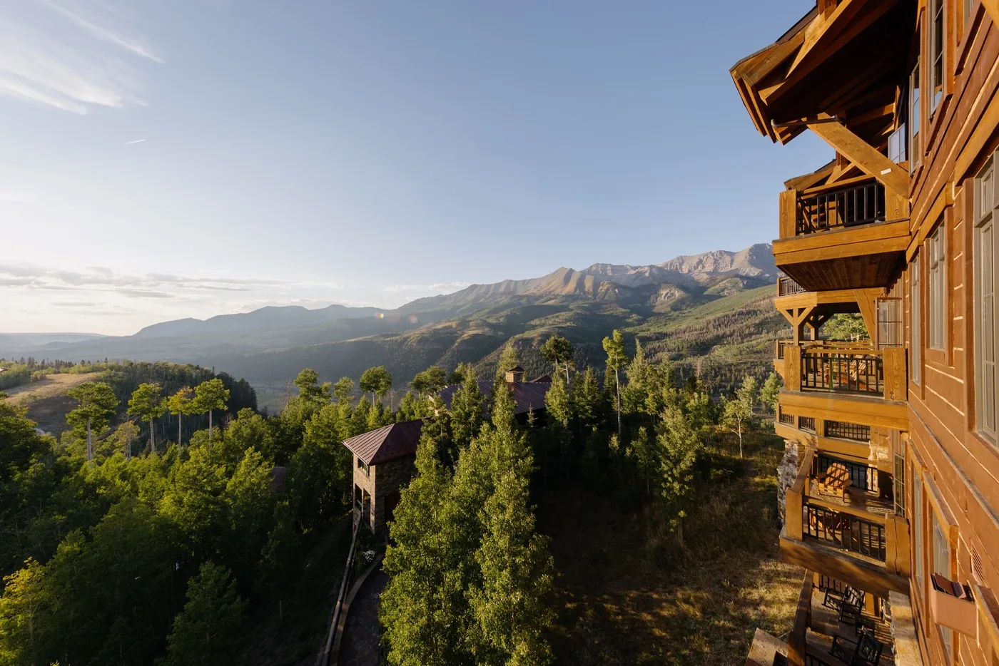 Un alojamiento de madera domina colinas boscosas y una cordillera bajo un cielo despejado.