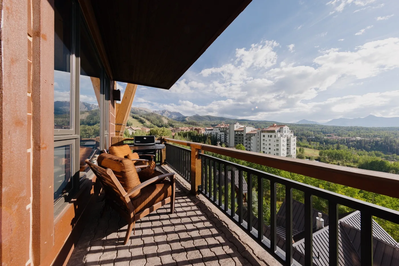 A balcony with outdoor seating overlooks buildings and green hills.