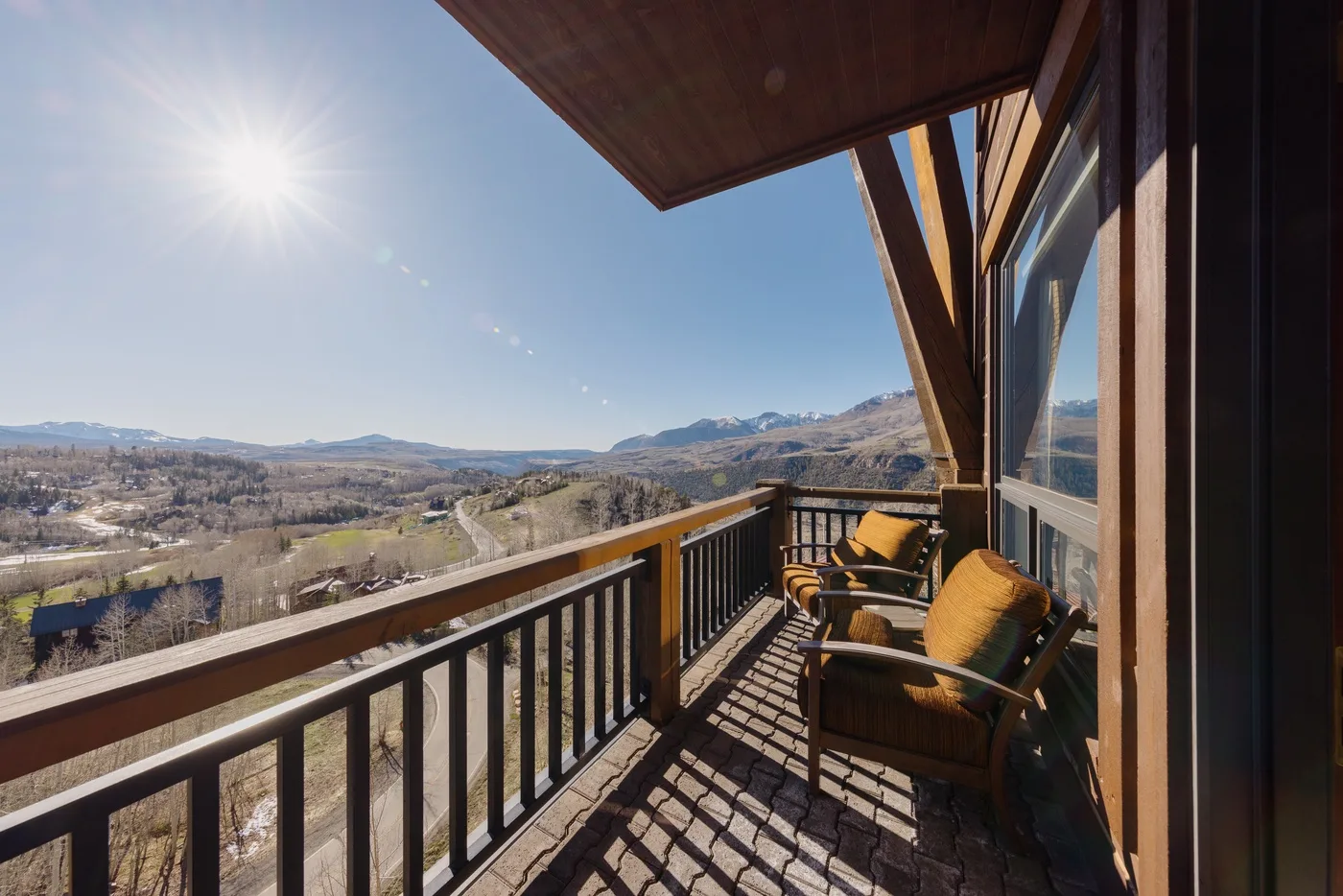 A sunny outdoor balcony overlooks a broad valley, with seating beside the railing.