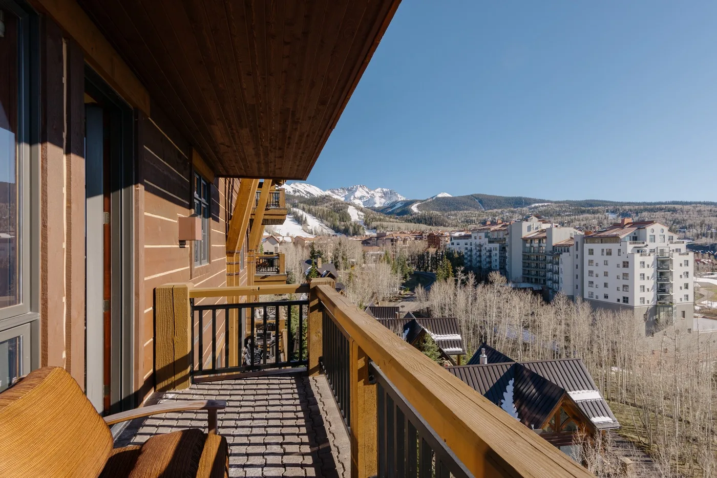 A balcony overlooks apartment buildings and snow-covered mountains beneath a clear sky.