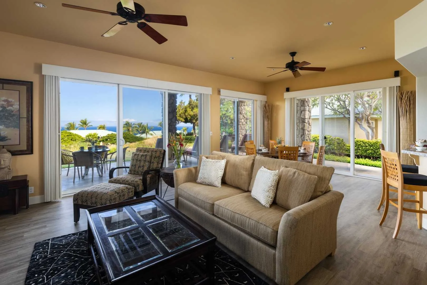 A bright living room with seating, a glass coffee table, and large windows overlooking greenery.