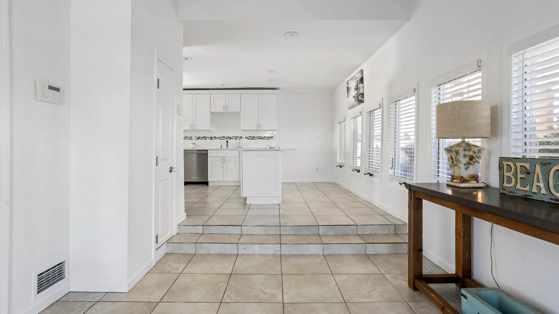 A bright kitchen opens onto a tiled living area with several windows.