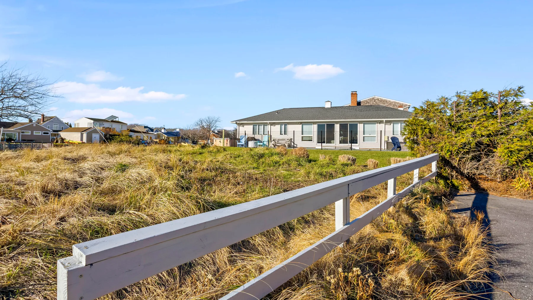 A house stands beyond a grassy lot beside a paved path and white railing.