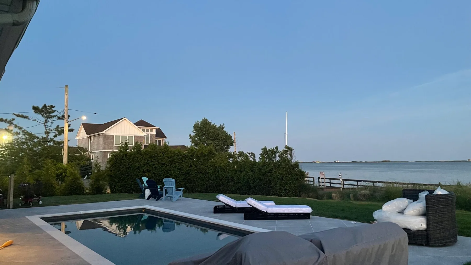 An outdoor pool beside a waterfront, with lounge seating and trees nearby.