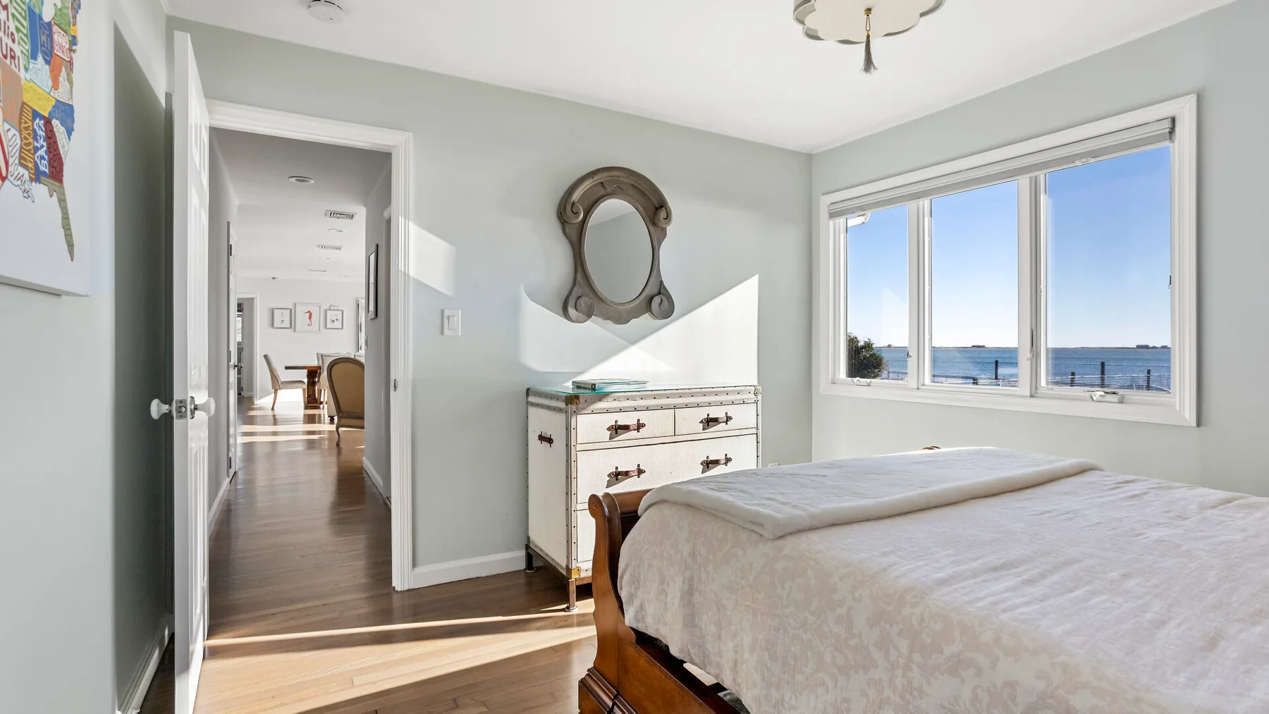 A bright bedroom with a bed, dresser, and a large window overlooking the water.