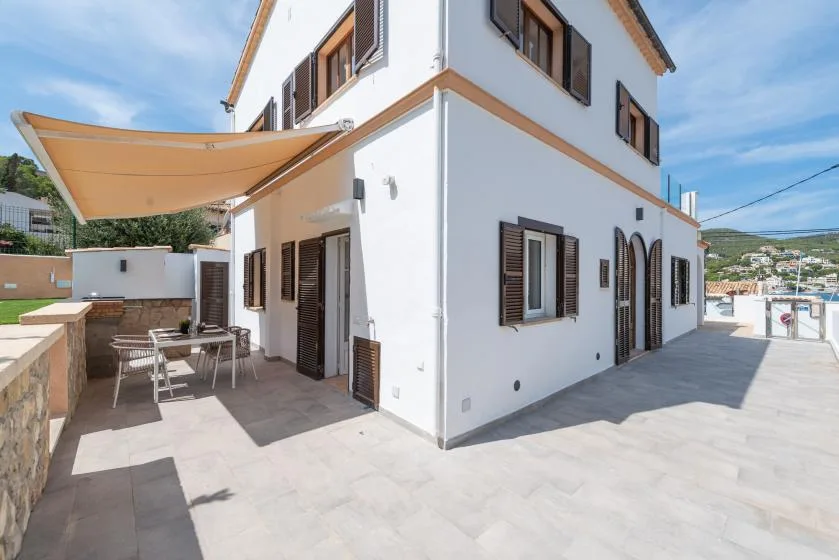 A white two-story house opens onto a paved patio with outdoor seating beneath a shade canopy.