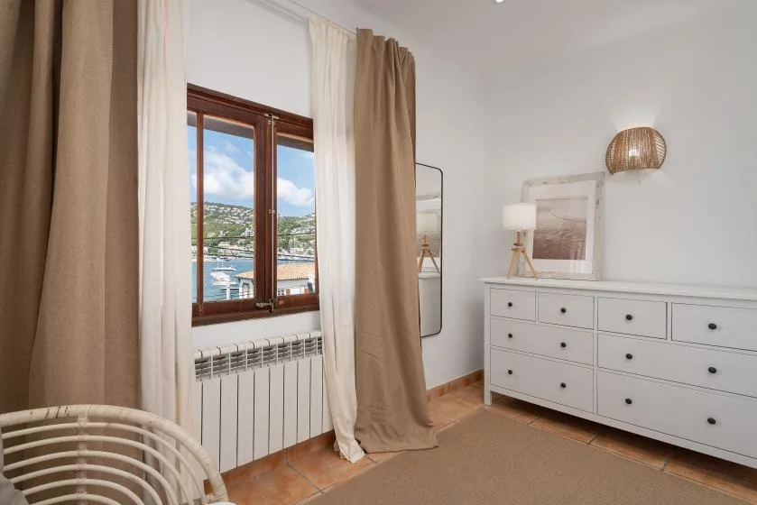 A bright bedroom with a window overlooking the water and a dresser.