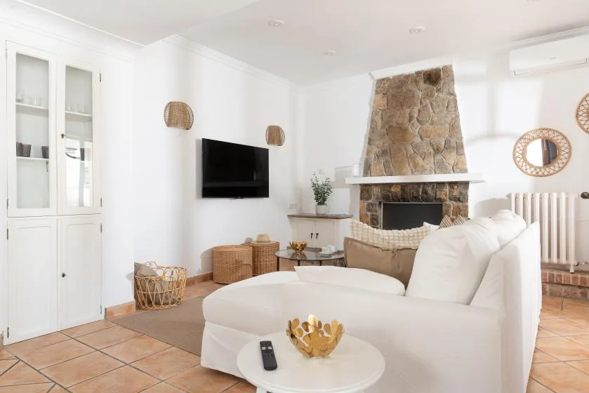 A bright living room with white seating, a wall-mounted television, and a stone fireplace.